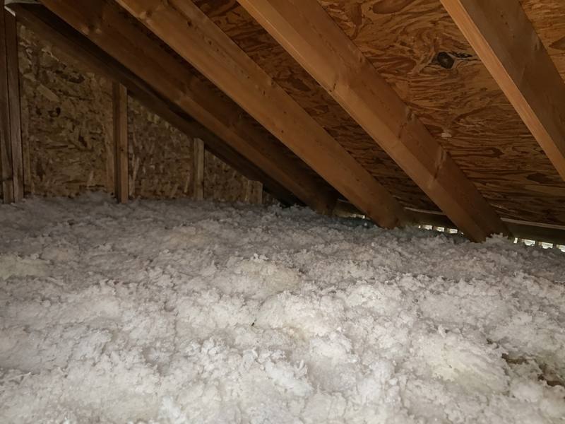 Attic floor covered in deep white blown-in insulation under wooden rafters and a plywood roof deck