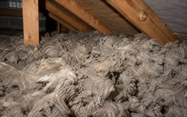 Old grey rock wool insulation piled loosely across an attic floor below the rafters