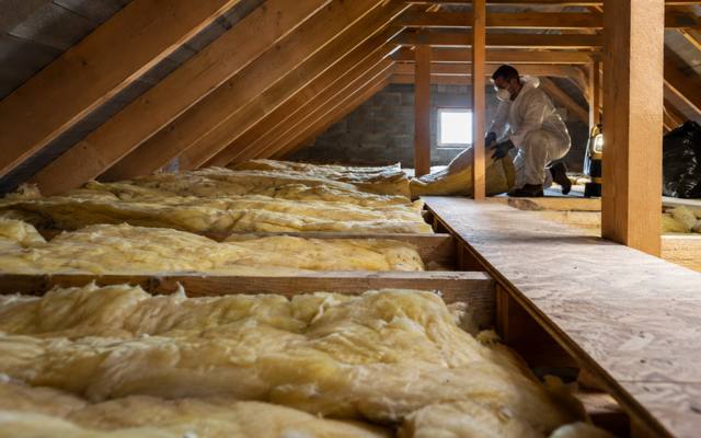 Worker in a protective suit handling yellow batts near the eaves of an attic beside a walkway
