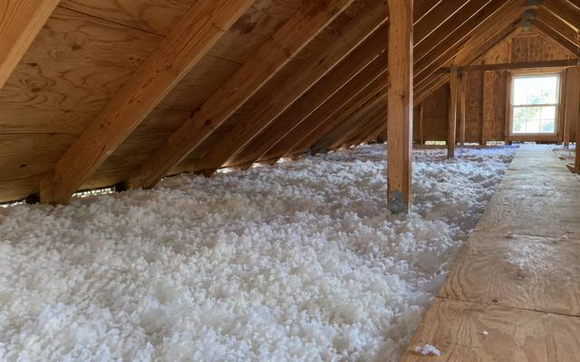 Fresh white blown-in insulation beside a plywood walkway in an attic with a gable window
